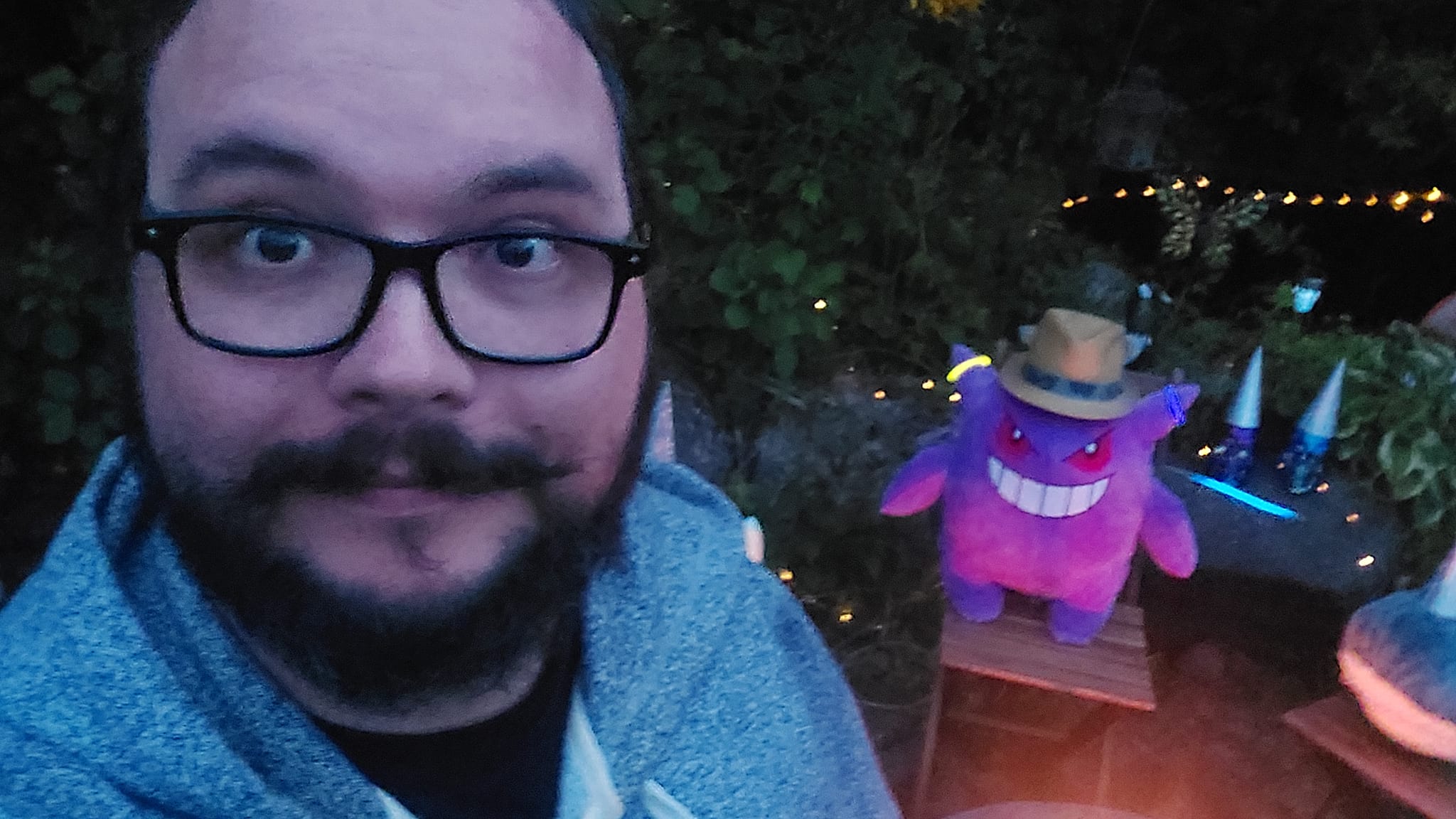 A man with facial hair stands in front of a stuffed animal campfire, Gengar is visible in the background